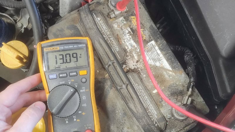 A professional mechanic testing car battery voltage using a digital multimeter in the engine bay under natural daylight.